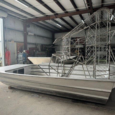 Unfinished aluminum boat inside a workshop, with a tall metal lattice framework mounted at the stern and an open bay door visible in the background.