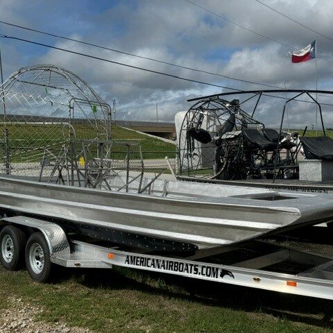 Two aluminum airboats on a dual-axle trailer in a grassy lot; open cockpit frames with protective cages, trailer labeled AmericanAirboats.com.