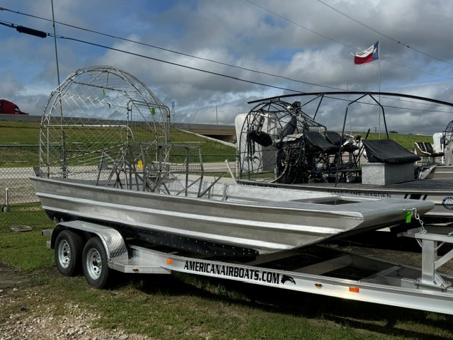 18x8_AirRanger Two aluminum airboats on a dual-axle trailer in a grassy lot; open cockpit frames with protective cages, trailer labeled AmericanAirboats.com.