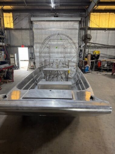 Metal boat hull under construction in a machine shop, with skeletal frame at the bow and metal panels along the sides, workshop tools visible in the background.