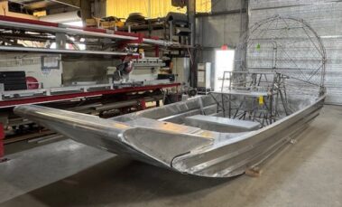 Unfinished aluminum boat hull rests on supports inside a workshop with metal shelving and equipment nearby.