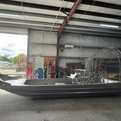 Aluminum airboat inside a workshop, long hull with a circular propeller cage at the rear and a metal frame seating area.