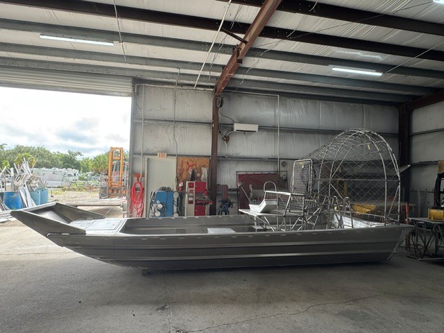20x8_side_profile Aluminum airboat inside a workshop, long hull with a circular propeller cage at the rear and a metal frame seating area.
