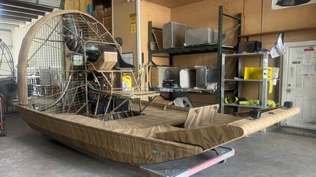 Airboat with a circular rear propeller cage, stored in a workshop with metal shelving and tool equipment nearby.