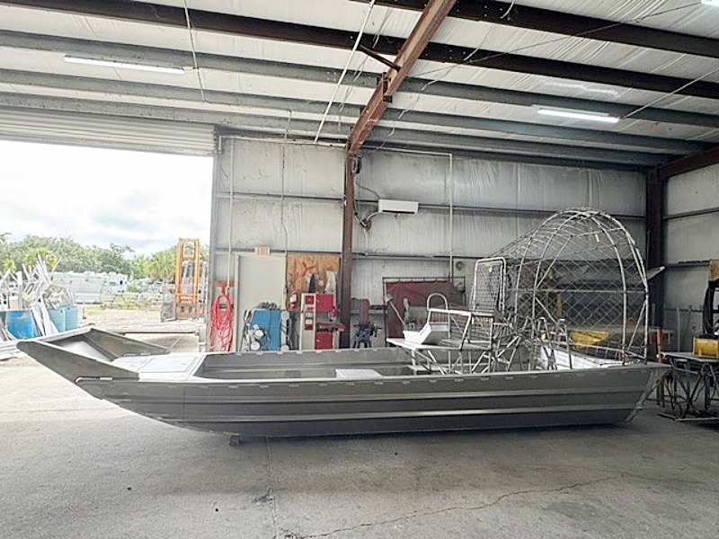 An aluminum airboat inside a metal workshop, with the propeller cage visible on the right and equipment in the background